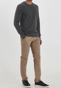 Hombre vistiendo una camiseta de manga larga gris oscuro, pantalones beige y zapatillas negras, de pie con las manos en los bolsillos contra un fondo neutro.