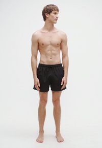 Juodi plaukimo šortai su elastiniu juosmeniu, su raišteliu. Modelis lieso sudėjimo, stovintis basas priešais paprastą baltą foną.