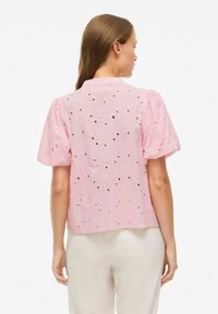 Blouse rose à manches bouffantes avec broderie, présentant des motifs floraux découpés. Tissu léger, conception courte et une finition texturée subtile.