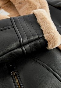 Sort faux læderjakke med en blød tan faux pelskrave. Har horisontal syning og en synlig guld lynlås detalje.