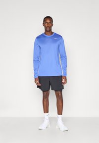 Blå langærmet træningsshirt med rund halsudskæring og logo, parret med sorte shorts. Båret med hvide sneakers og sokker.