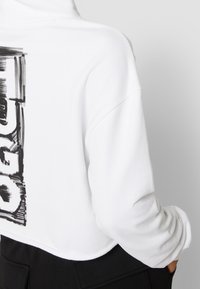 White cropped hoodie with black graphic text on the back, worn with black pants, showing folded sleeve and hand in pocket.