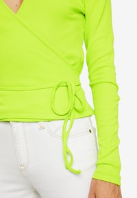 Neongrön ribbad wrap-overdel med långa ärmar och en knytning vid midjan, tillsammans med vita jeans med framficka.