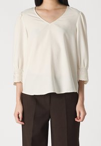 Blusa color crema con scollo a V, maniche a sbuffo e decorazioni ricamate ai polsini. Tessuto morbido e vestibilità rilassata, abbinata a pantaloni marroni.