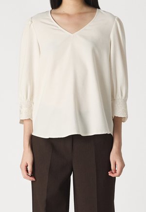 Cremefarbige Bluse mit V-Ausschnitt, Puffärmeln und bestickten Akzenten am Bündchen. Weicher Stoff und entspannter Schnitt, kombiniert mit braunen Hosen.