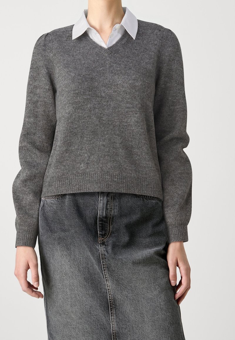 Personne portant un pull gris à col en V par-dessus une chemise blanche à col, et une jupe en denim noire taille haute, debout devant un fond uni.