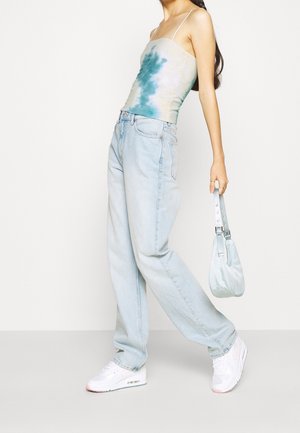 Femme portant un jean large bleu clair, des baskets blanches, un débardeur tie-dye bleu-vert, tenant un petit sac à main bleu clair à ses côtés.