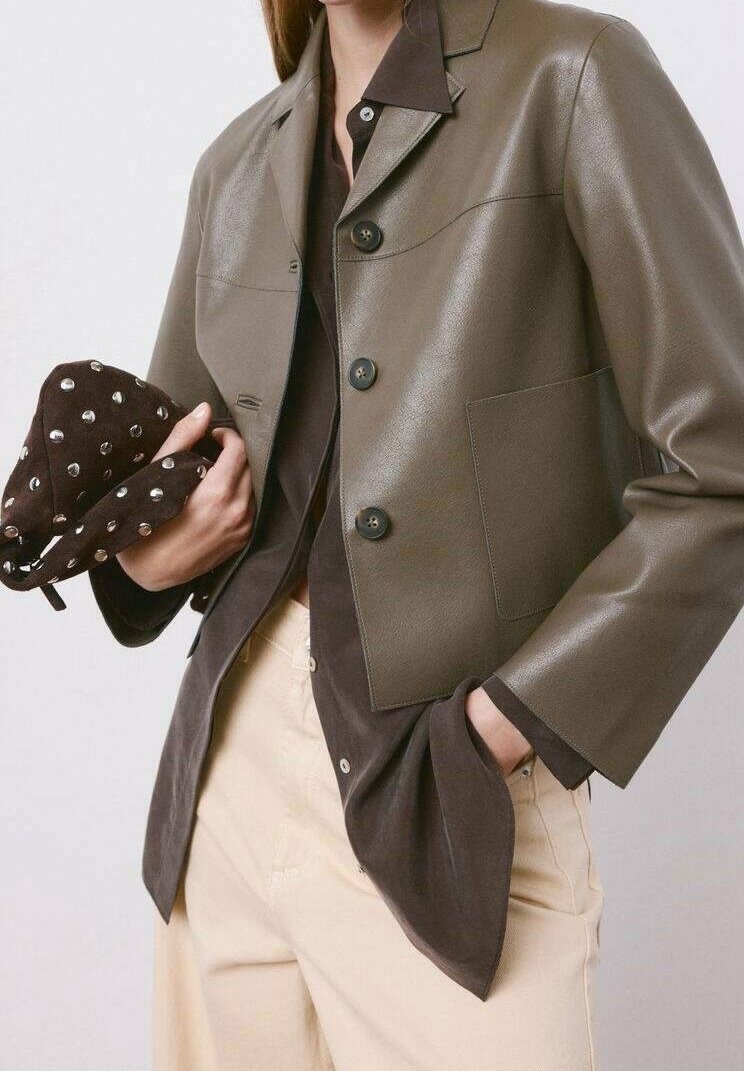 Personne portant une veste en cuir olive courte par-dessus une chemise sombre et un pantalon beige, tenant un chapeau marron foncé clouté d'une main.