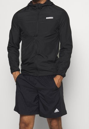 Mann trägt einen schwarzen Jack & Jones Reißverschluss-Hoodie und schwarze Adidas-Shorts mit Händen in den Taschen vor einem einfarbigen Hintergrund.