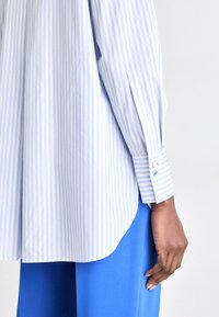 Paže se středním odstínem pleti, která má na sobě modrobílý proužkovaný dlouhý rukáv a jasně modré kalhoty na jednobarevném pozadí.