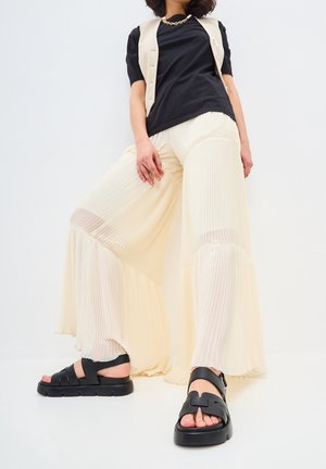 Kermansävyiset laskostetut leveälahkeiset housut, joissa on läpikuultavia yksityiskohtia ja hentoinen helma, yhdistettynä mustiin paksupohjaisiin sandaaleihin. Yläosana on musta vartalonmyötäinen t-paita.