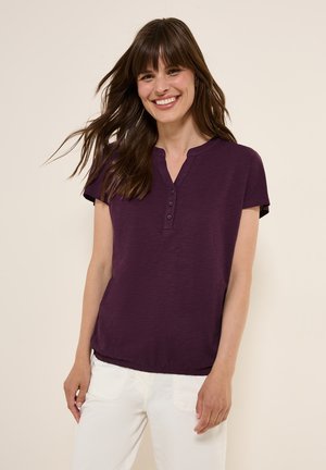 Top corto viola a maniche corte con scollo henley e pattina di bottoni; tessuto leggero con una vestibilità rilassata, abbinato a pantaloni bianchi.