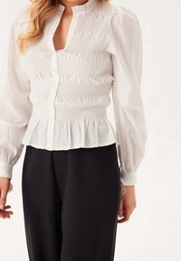Weiße, gerüschte Bluse mit hohem Kragen, Knopfleiste und ausgestelltem Saum. Lange, gepuffte Ärmel mit elastischen Bündchen.