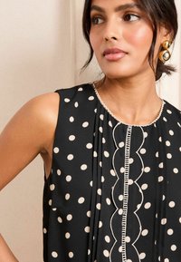 Mustakatehena ilma soe blouse suurte kreemjate täppidega, volditud disain, laineline kaelus kontrastse äärega, sile kangast tekstuur.