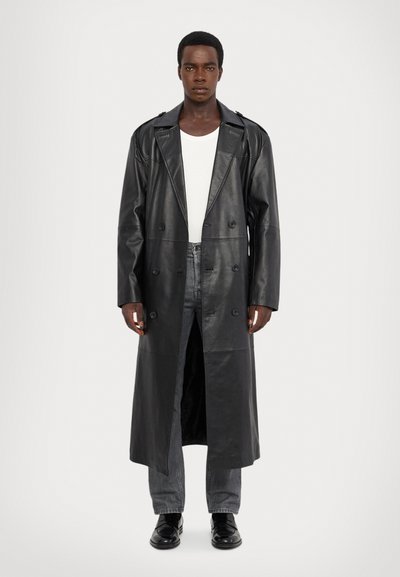 Svart läder dubbelknäppt trench coat med breda slag, skräddarsydd passform och lång siluett. Bärs över en vit T-shirt och grå jeans.