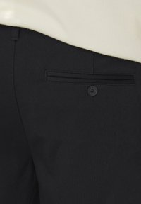 Pantaloni neri con una singola tasca posteriore con chiusura a bottone, design slim e texture liscia. Il capo superiore è di colore chiaro.
