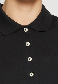 Czarna koszulka polo z teksturowanej tkaniny, z klasycznym kołnierzykiem i pięcioma kontrastowymi białymi guzikami, prezentująca schludny i dopasowany krój.
