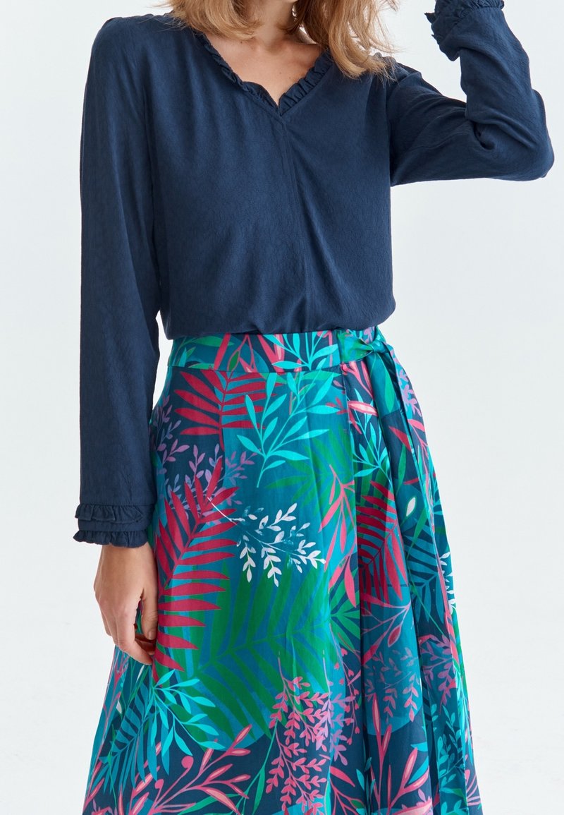 Blusa blu navy a maniche lunghe con scollatura arricciata abbinata a una lunga gonna maxi colorata e decorata con foglie tropicali nei toni del rosa e del verde.