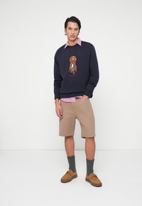 Mornarska dukserica z vezenim vzorcem psa, v kombinaciji s svetlo roza majico s kragnico, bež teksturiranimi kratkimi hlačami, sivimi nogavicami in rjavimi čevlji.