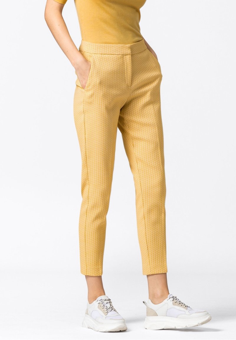 Pantalon jaune à motifs avec une coupe slim, doté de poches et d'une texture lisse, associé à des baskets blanches et grises.