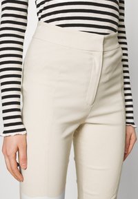 Béžové kalhoty na míru s vysokým pasem, se zaoblenými předními švy a hladkou texturou. Nosené s černobílým pruhovaným tričkem s dlouhým rukávem.