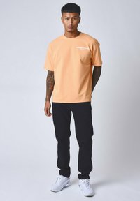 Camiseta de algodón de color durazno con mangas cortas y un logotipo de texto blanco en el pecho, combinada con pantalones negros y zapatillas blancas.