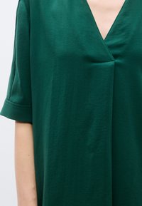 Haut vert à manches courtes avec un décolleté en V, présentant un design simple, une texture douce et un léger pli près de la zone de la poitrine.