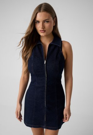 Jeune femme aux longs cheveux portant une robe en jean bleu foncé sans manches avec une fermeture éclair sur le devant, debout devant un fond uni.
