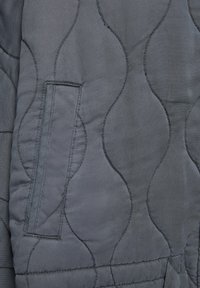 Prešívaná sivá tkanina s vlnitým vzorom prešívania a vertikálnym vreckom s lemom.