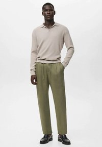 Camisa polo de manga larga en beige con ribetes, combinada con pantalones en verde oliva que cuentan con una cintura ajustable y bolsillos laterales. Unos zapatos negros completan el atuendo.