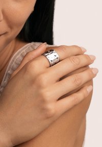 Ampio anello d'argento con una texture martellata, caratterizzato da un interno liscio e una forma leggermente curva, indossato su un dito. Colore della pelle visibile.