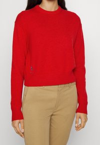 Nainen, jolla on päällään kirkkaan punainen pitkähihaisen neule, jossa on pyöreä pääntie ja beiget housut. Neuleen helman lähellä on pieni tummansininen logo.