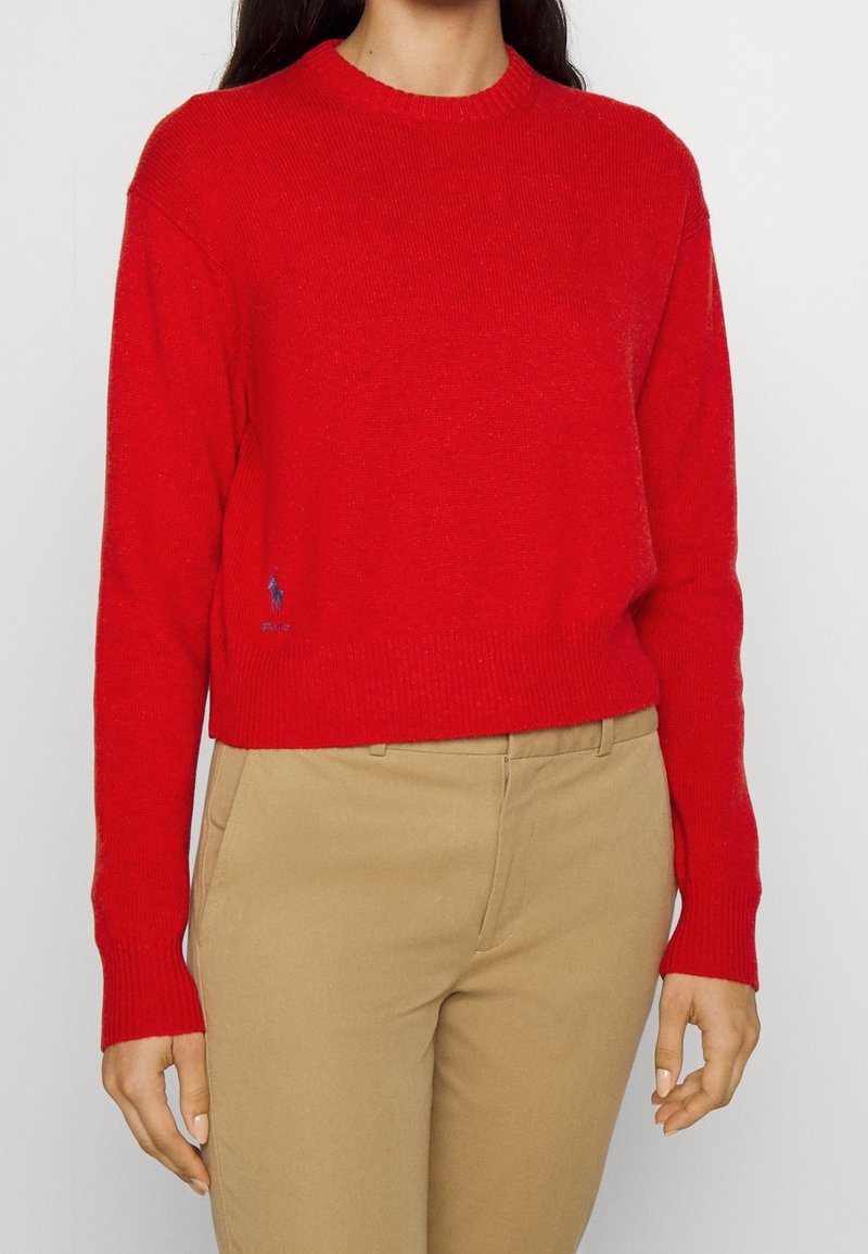 Nainen, jolla on päällään kirkkaan punainen pitkähihaisen neule, jossa on pyöreä pääntie ja beiget housut. Neuleen helman lähellä on pieni tummansininen logo.