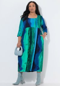 Lang kjole med et livligt blåt og grønt bølgemønster, rund halsudskæring, trekvartlange ærmer og lommer, kombineret med en lyseblå håndtaske.