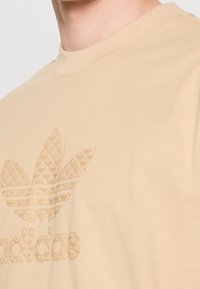 Camiseta de algodón beige con un logo de adidas texturizado y en relieve, y un acento de tres hojas. Cuello redondo, mangas cortas y un corte relajado.