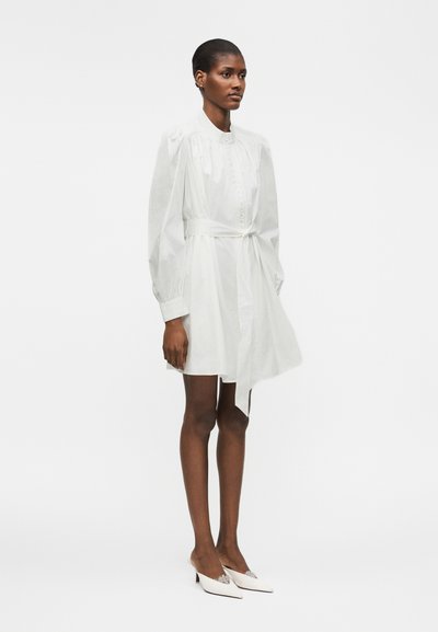 Γυναίκα που φοράει ένα λευκό μίνι φόρεμα με μακριά μανίκια και ζώνη στη μέση, με λευκά μυτερά ψηλοτάκουνα, στέκεται μπροστά σε απλό φόντο.