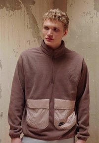 Hnedý fleecový pulóver s vysokým golierom, polosťahovacím zipsom a dvoma béžovými prednými vreckami. Obsahuje logo Nike na pravom vrecku. Mäkka textúra.