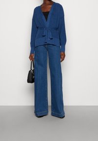 Blå stickad cardigan med knytmidja, tillsammans med vida blå jeans. Outfitten inkluderar en svart handväska för kontrast.