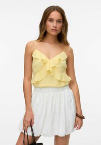 Geel chiffon top met ruffle-ontwerp en dunne schouderbandjes, gecombineerd met een witte, textuurrok. Zwarte handtas en bruine armband accentueren de outfit.