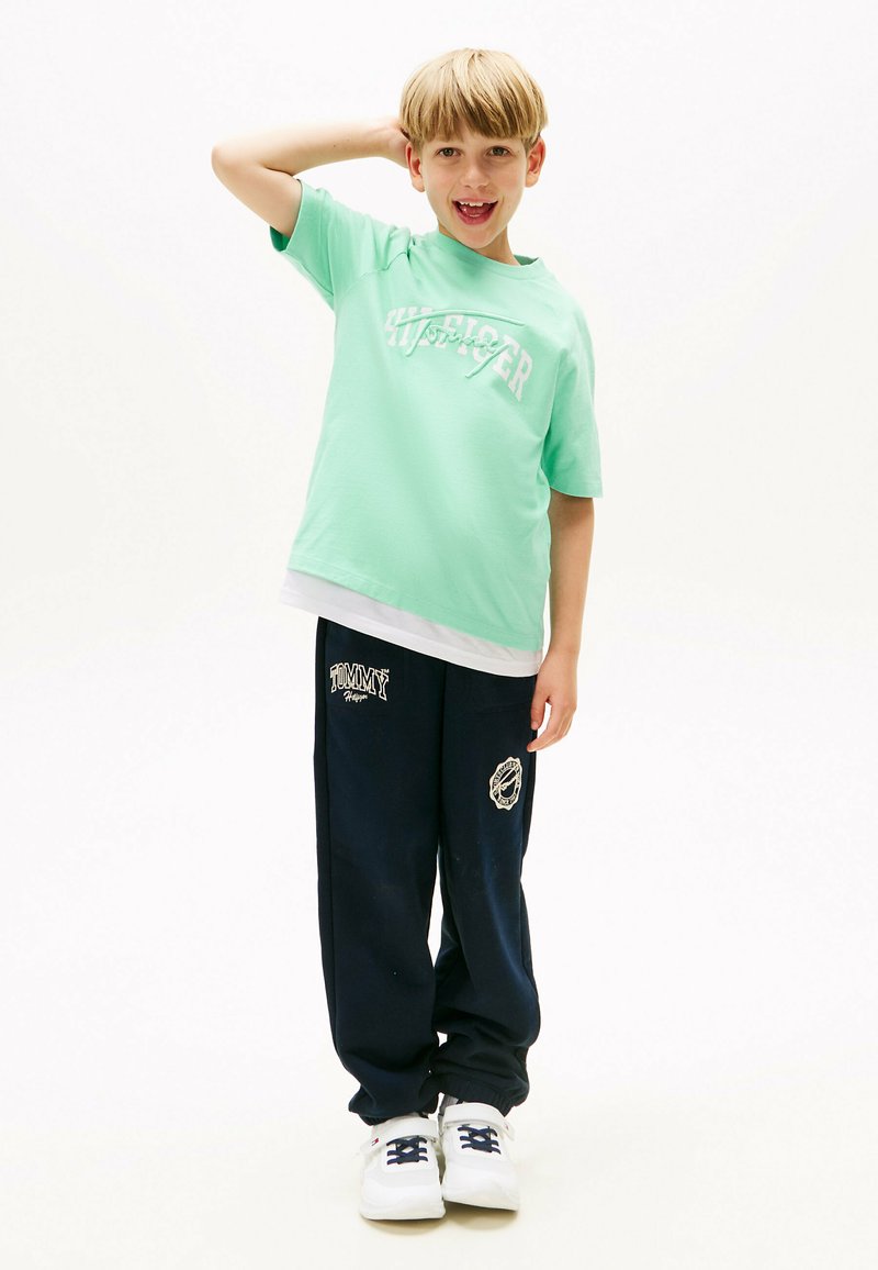T-shirt verde in cotone con logo bianco, abbinato a pantaloni jogger blu navy con accenti e grafiche bianche. Sneakers bianche con lacci scuri.