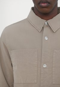 Close-up of a person wearing a beige shirt jacket with two large chest pockets and three silver snap buttons down the front.