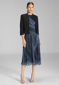 Czarna krótka kurtka na granatowej sukience z teksturowanym, spiralingowym wzorem. Sukienka ma zwiewny dół i jest zestawiona z sandałkami na obcasie w kolorze srebrnym.