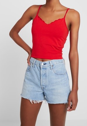 Mujer con top corto rojo sin mangas y pantalones cortos vaqueros de tiro alto en azul claro con dobladillos deshilachados, de pie con una mano en la cadera.