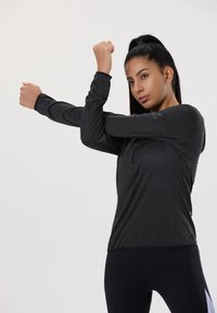 Schwarzes, sportliches Langarmshirt aus strukturiertem Stoff, mit hohem Kragen und taillierter Passform, getragen beim Dehnen der Arme.