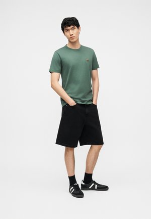 Joven de pie con las manos en los bolsillos, vistiendo una camiseta verde, pantalones cortos negros hasta la rodilla, calcetines negros y zapatillas negras con rayas blancas.
