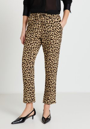 Donna che indossa pantaloni a stampa leopardata e scarpe nere con tacco kitten a punta e cinturino sul tallone, con una mano in tasca, in piedi su un pavimento chiaro.