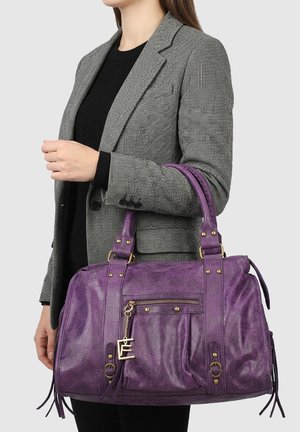 Vrouw in een grijze blazer houdt een grote paarse leren handtas vast met een voorvak met rits en een decoratieve gouden hanger.