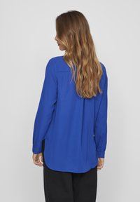 Chemise bleue à manches longues en tissu léger, avec un léger éclat, un ourlet incurvé et des poignets à boutons. Comprend une empiècement au dos.