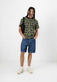Svart kortermet skjorte med flerfargede mønstre, kombinert med blå denimshorts og beige sneakers, og en svart skulderveske.