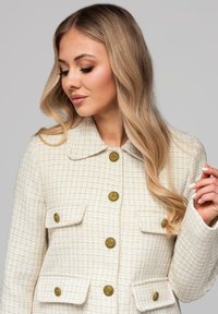 Kremowa marynarka z tweedu o fakturowanym wykończeniu, z dużym kołnierzem, złotymi guzikami i dwoma przednimi kieszeniami. Wyraźny wzór w kratę na całej powierzchni.